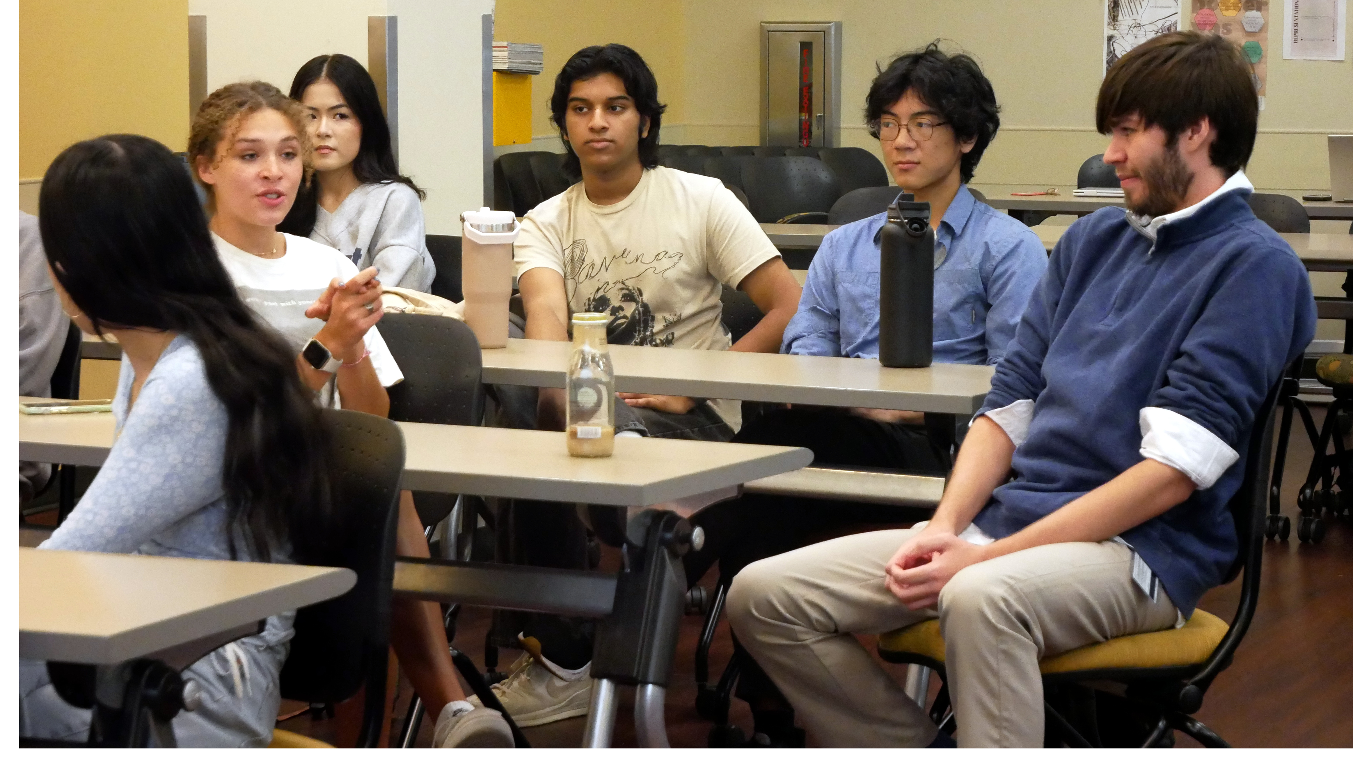 A diverse group of college students sits in a classroom, engaged in a discussion. One young woman in a white t-shirt is speaking, while others attentively listen. Notebooks, drinks, and water bottles are visible on the desks.
